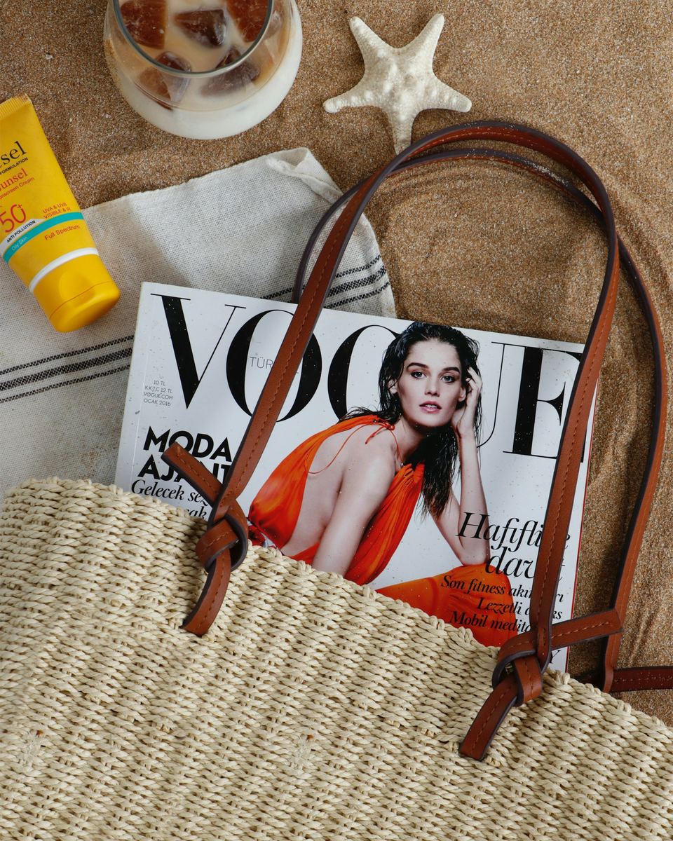 A straw tote bag with sunscreen and a magazine on a beach towel