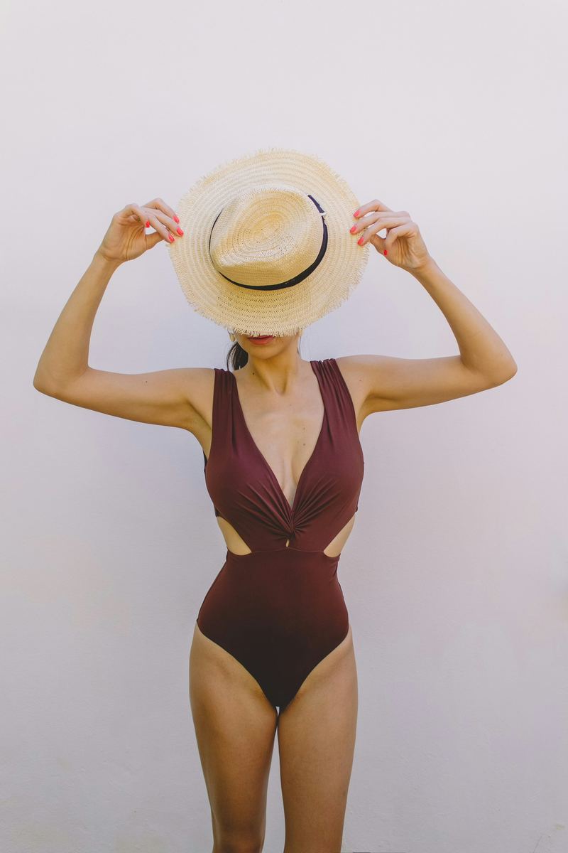Woman in a brown cutout one-piece swimsuit holding a straw hat